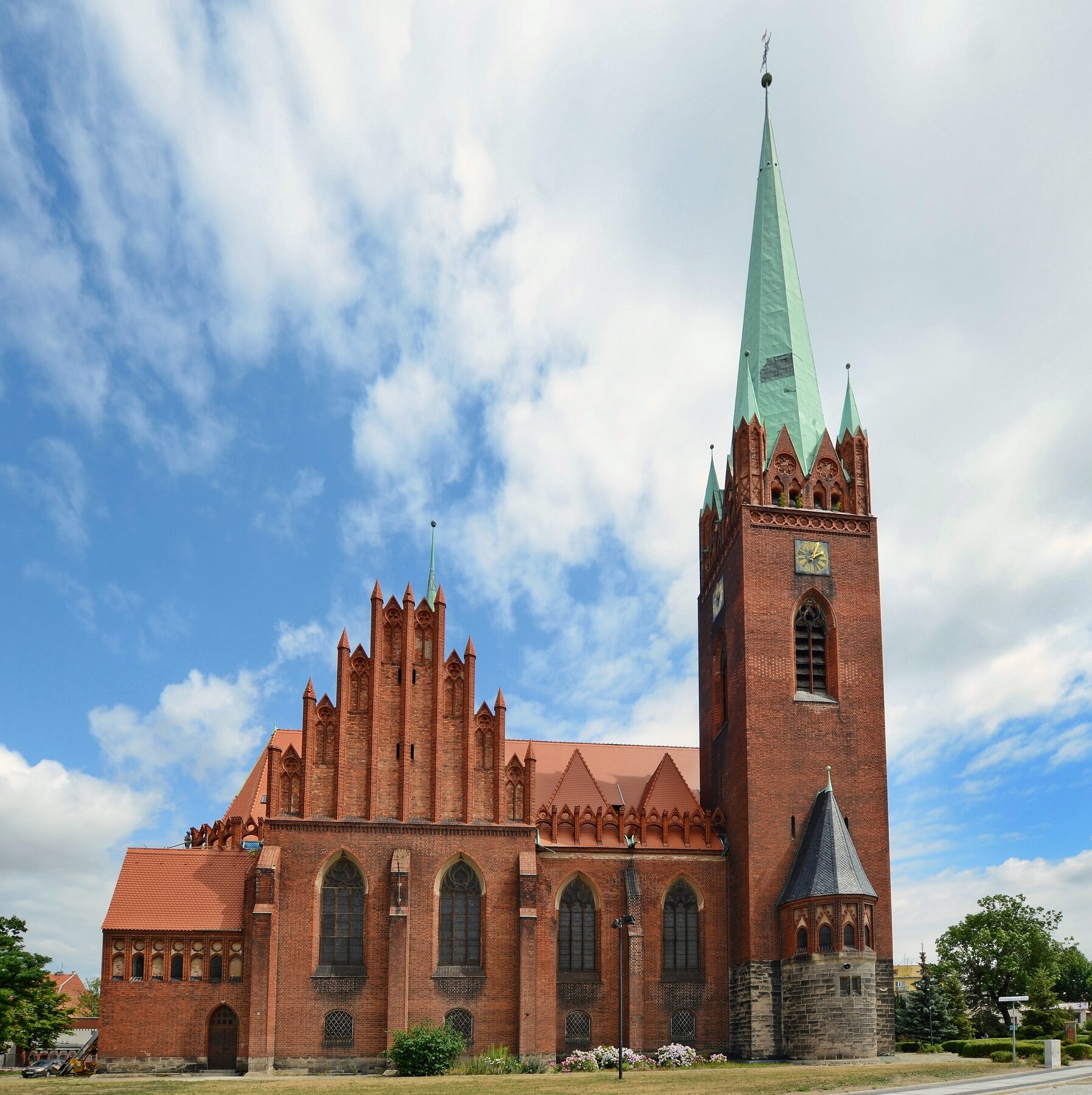 Torre con aguja verde y muros de ladrillo rojo de la iglesia de San Jacinto de Legnica, en Polonia, bajo un cielo con nubes
