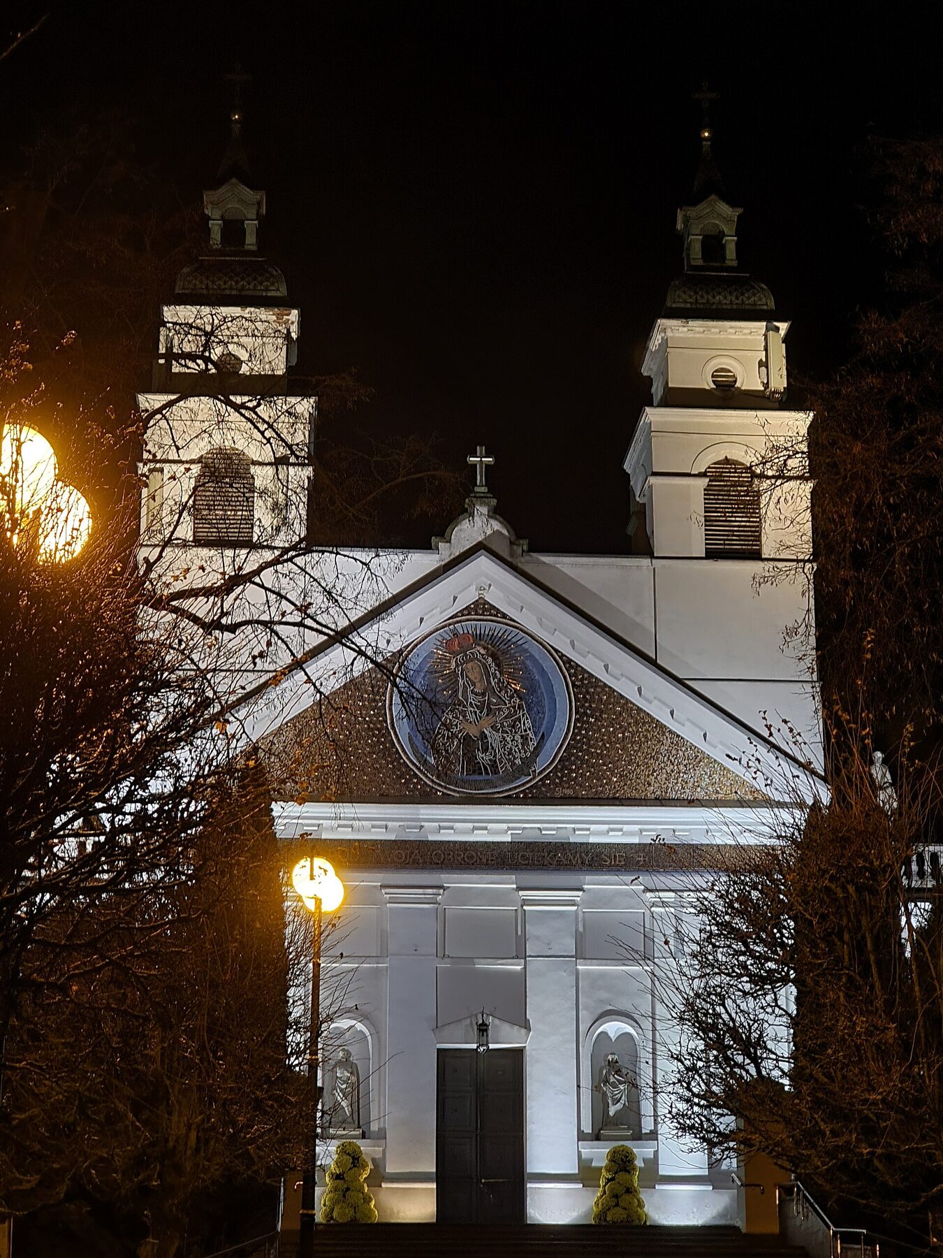 Fachada de la colegiata de San Antonio de Sokółka, con dos torres y una imagen circular sobre la puerta, iluminada de noche